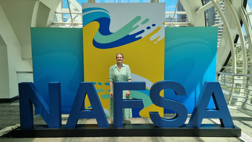 Person standing behind large blue 'NAFSA' letters, with colorful abstract panels and natural light in the background.